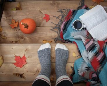 A cozy image of feet with socks on, in a fall setting, with an open book and coffee.