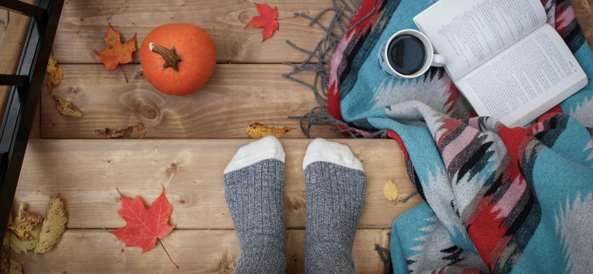 A cozy image of feet with socks on, in a fall setting, with an open book and coffee.