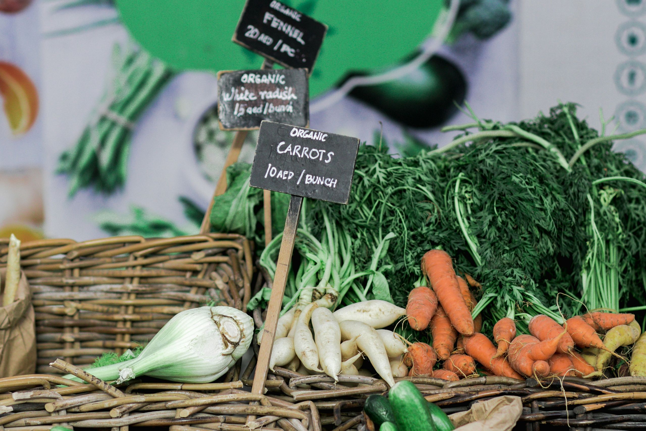 Carrots with green tops in a basket on the right. on the left, a bulb of fennel is in another basket. Chalkboard signs have descriptions of items and prices.