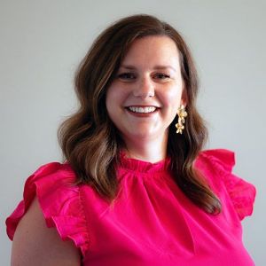 Mackenzie Pickerrell in a bright pink blouse with gold earrings against a gray backdrop.