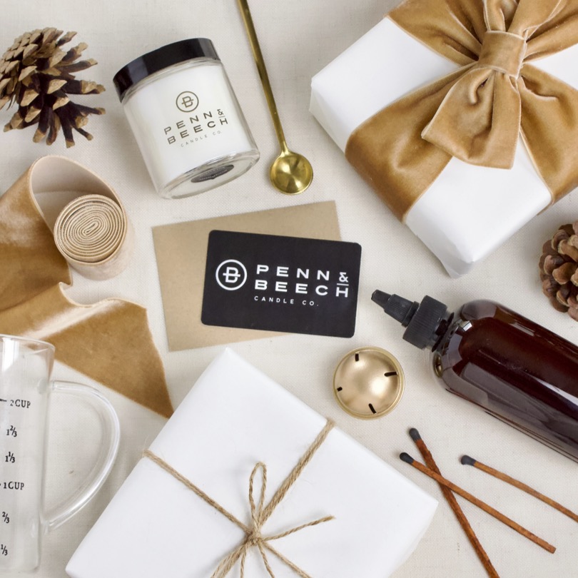 Penn & Beech Gift Card laying among white gifts wrapped with tan ribbon, pinecones and candle making supplies.