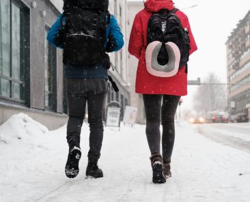 Two people walking on a snowy city street with backpacks. The one on the left has a blue coat, the right has red.