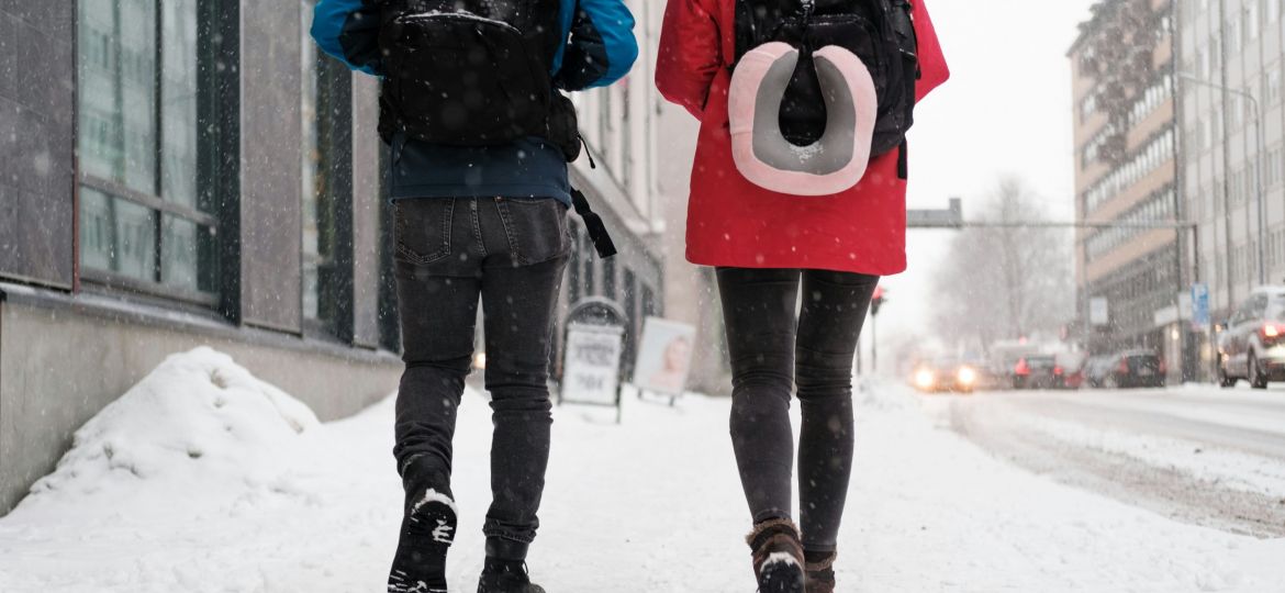 Two people walking on a snowy city street with backpacks. The one on the left has a blue coat, the right has red.