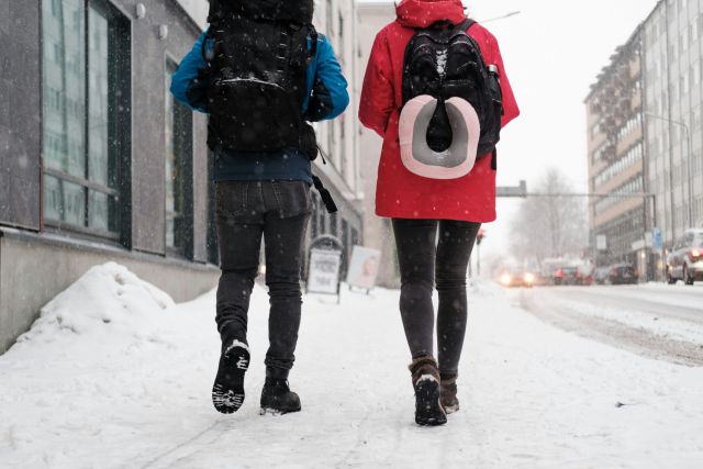 Two people walking on a snowy city street with backpacks. The one on the left has a blue coat, the right has red.