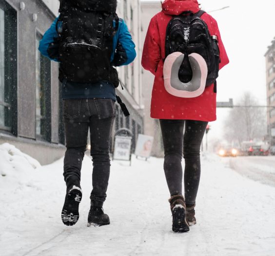 Two people walking on a snowy city street with backpacks. The one on the left has a blue coat, the right has red.