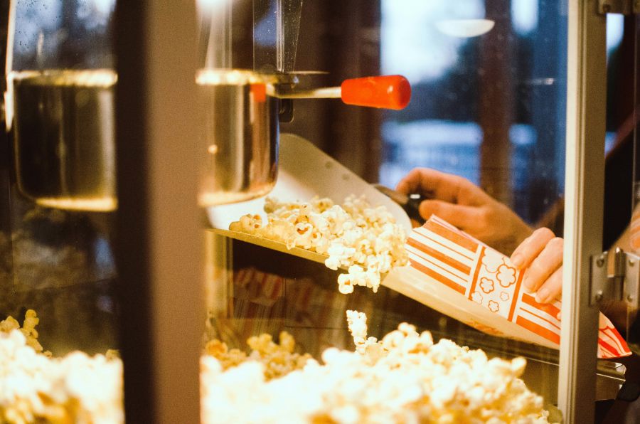 meg-boulden-A0FgI6AFjng-unsplash Close up image of hands scooping popcorn into a bag from a commercial machine