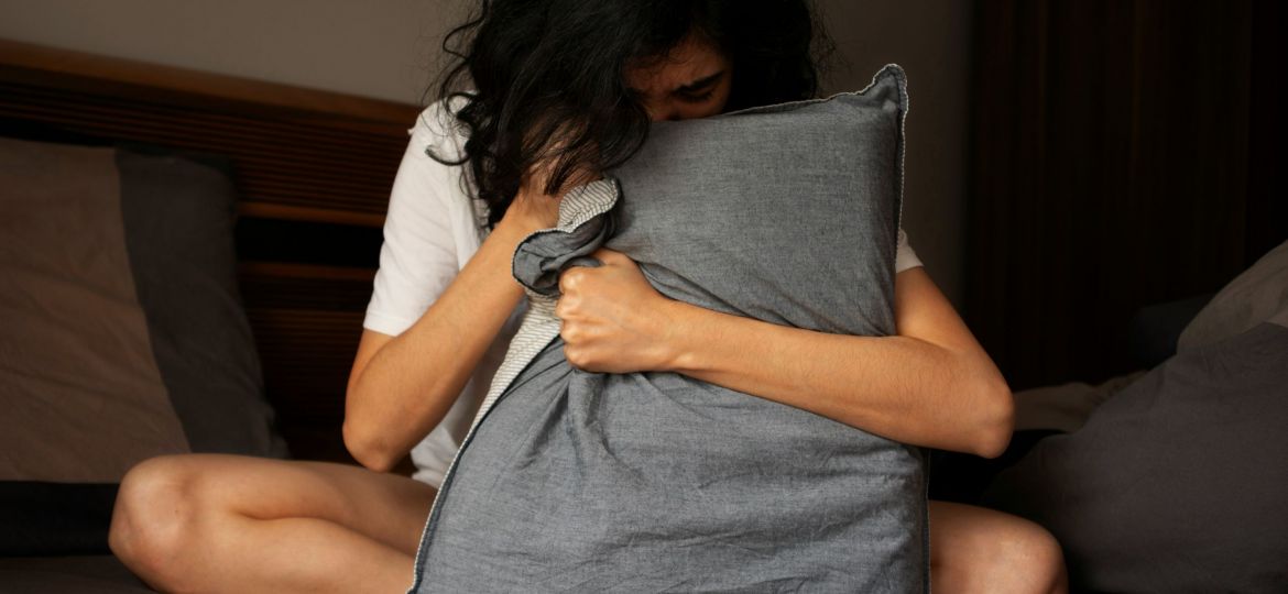 Woman with dark hair in white t-shirt gripping a gray pillow with her face buried in it.