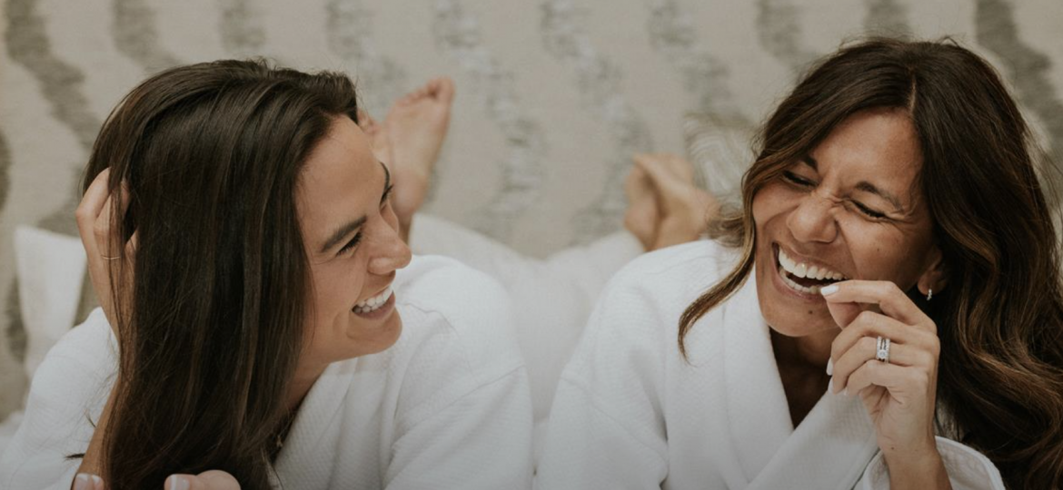 Two people laughing in a bed wearing robes.