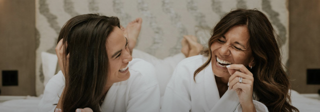Two people laughing in a bed wearing robes.