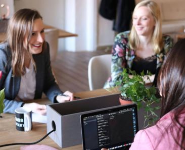 Three women talking in an office space, discussing business.