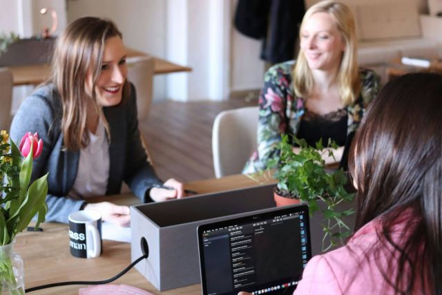 Three women talking in an office space, discussing business.